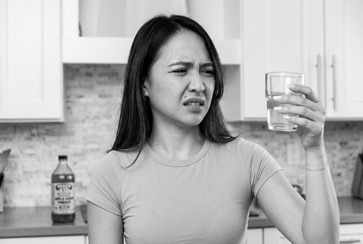 A woman grimacing after drinking apple cider vinegar, a bottle of it on the bench behind her